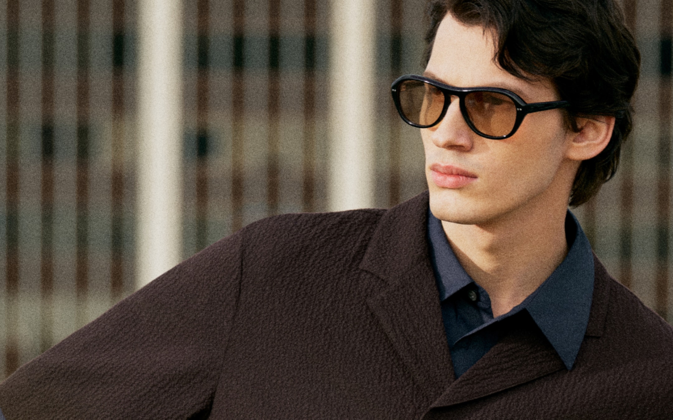 Man wearing dark sunglasses and a textured brown coat over a blue shirt, gazing to the side with a blurred building in the background.