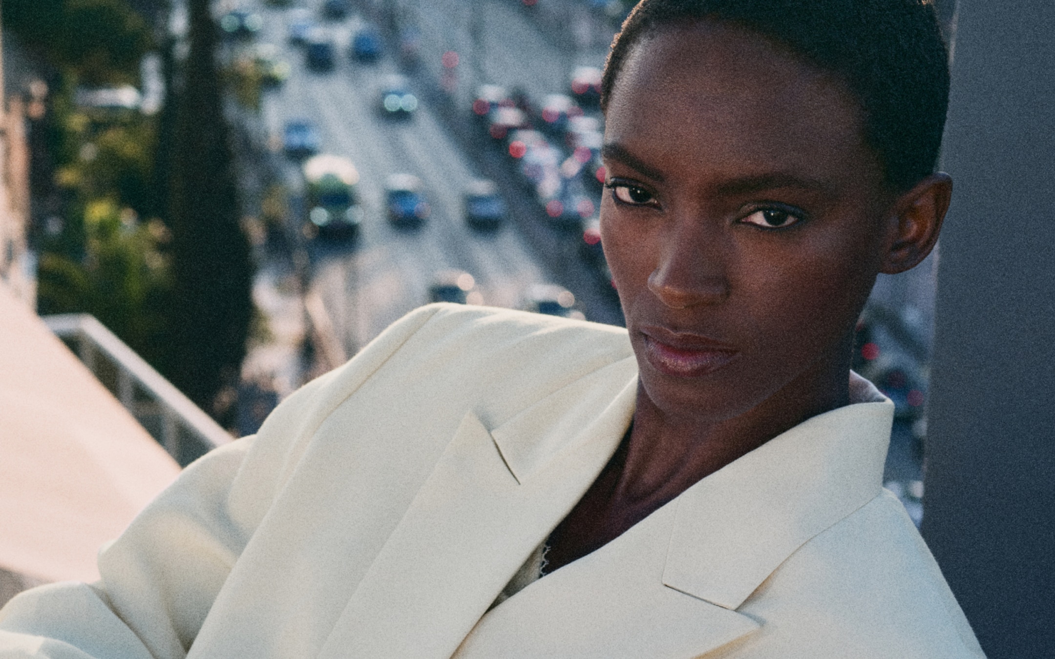 Person in a light-colored blazer looks confidently at the camera, with a blurred urban background and traffic below.