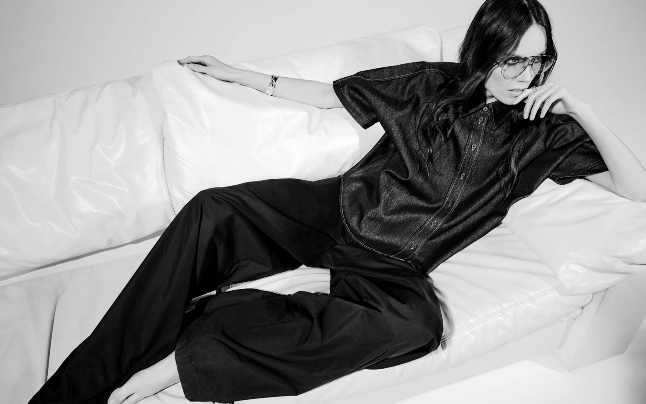 Person lounging on a white couch, wearing sunglasses, a dark short-sleeve shirt, and wide-leg pants, with a relaxed pose. Black and white image.