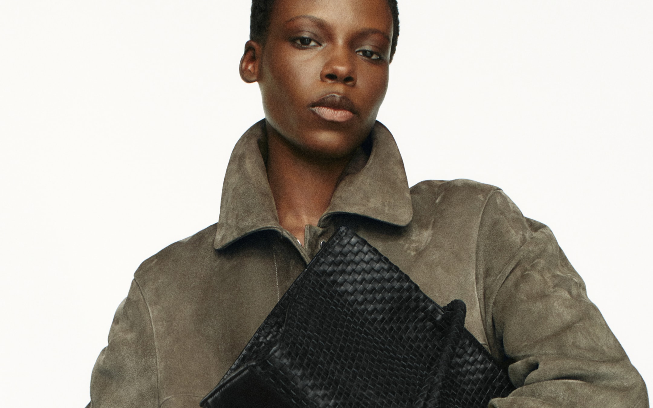 Close-up of a person in a suede coat holding a black woven leather clutch, neutral expression.