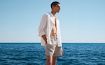 Placeholder for: Man in a white shirt and striped shorts stands by the ocean under a clear blue sky.