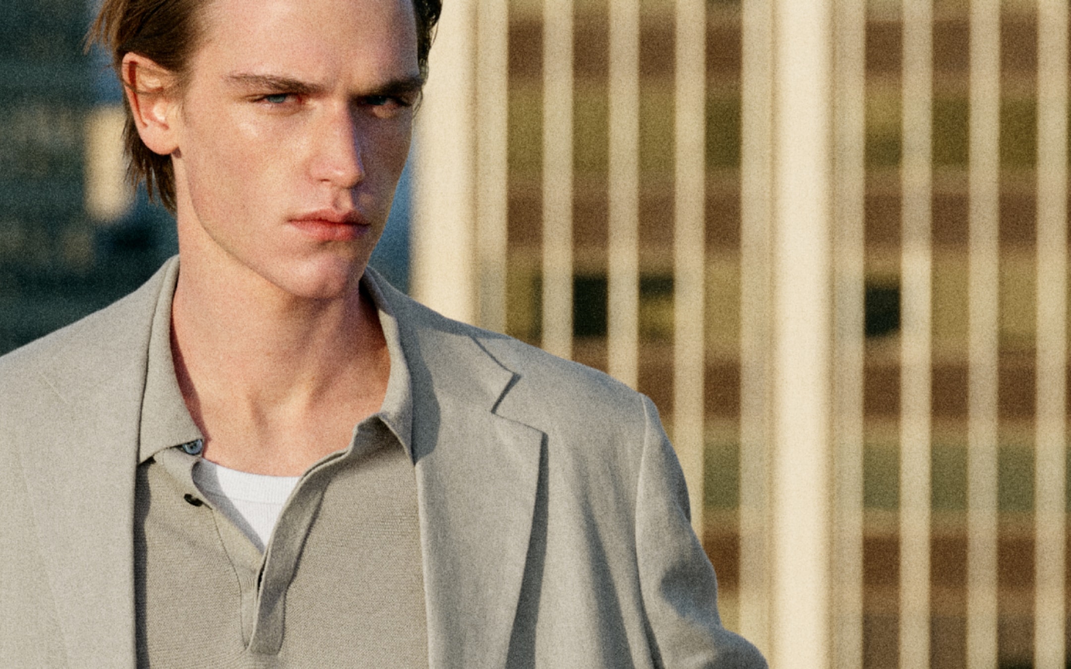 A person with short hair wearing a light gray blazer and polo shirt, standing outdoors with a blurred building in the background.
