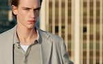 Placeholder for: A person with short hair wearing a light gray blazer and polo shirt, standing outdoors with a blurred building in the background.