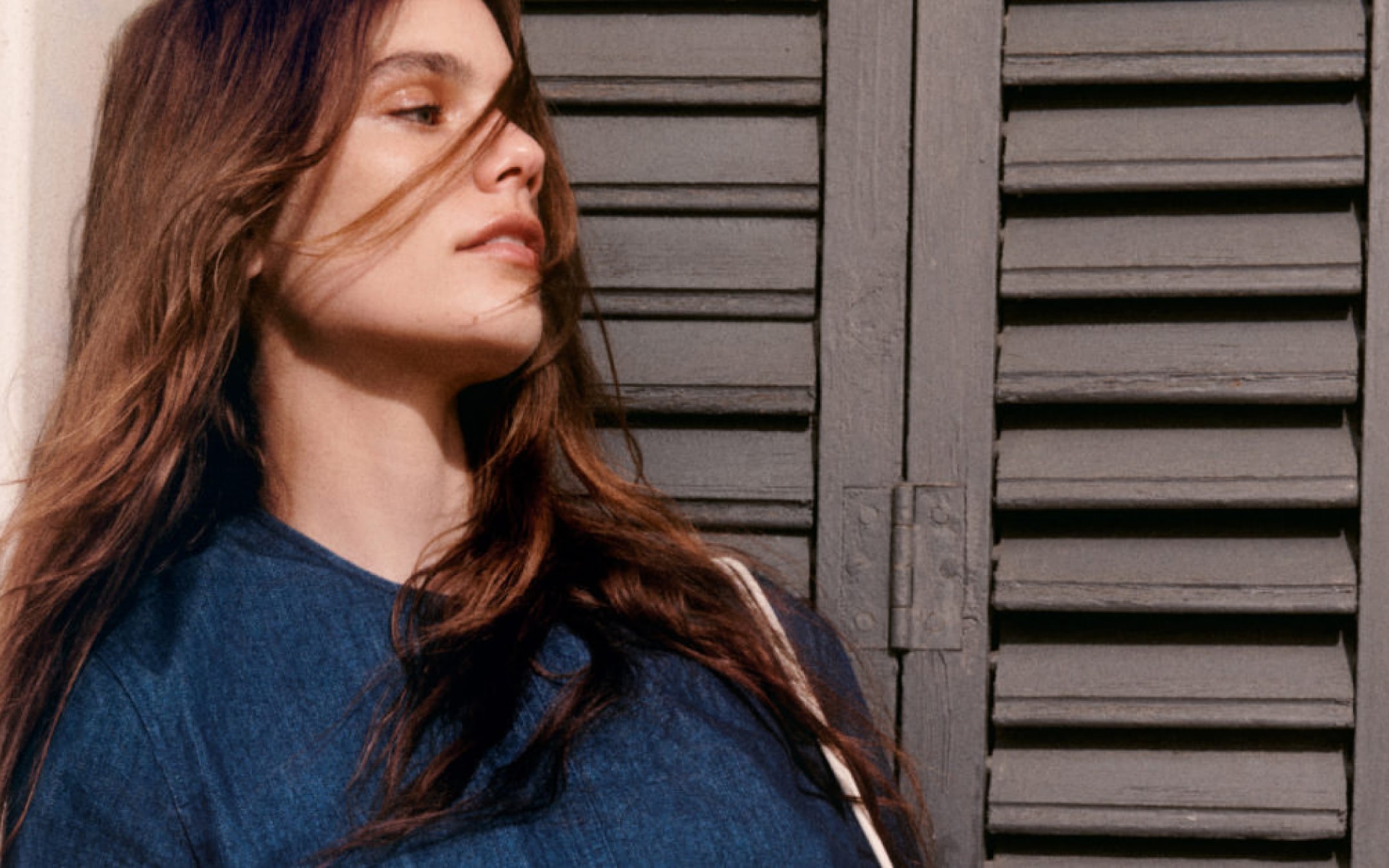Woman with long brown hair wearing a denim top, standing in front of a gray metal shutter, looking thoughtfully to the side.