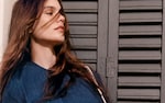 Placeholder for: Woman with long brown hair wearing a denim top, standing in front of a gray metal shutter, looking thoughtfully to the side.