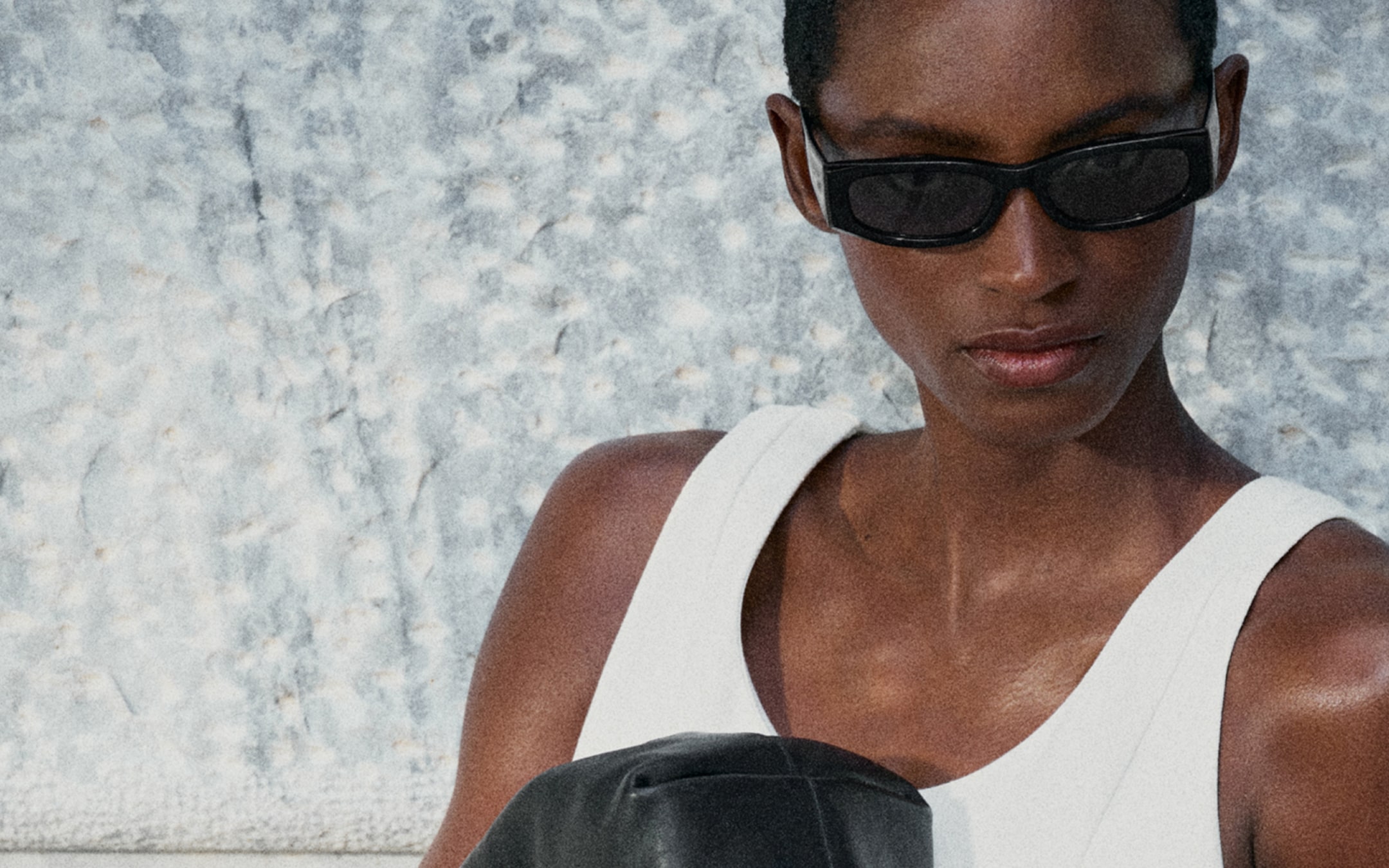Person in sunglasses and a white top in front of a textured wall, holding a black object, gazing downward.