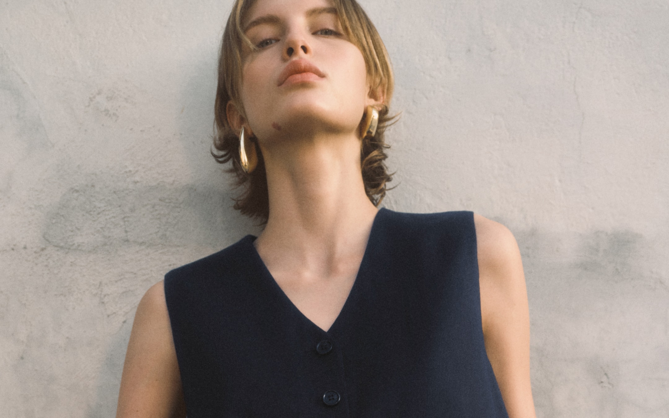 Model leaning against the wall. She's wearing golden hoop earrings and is wearing a black top.