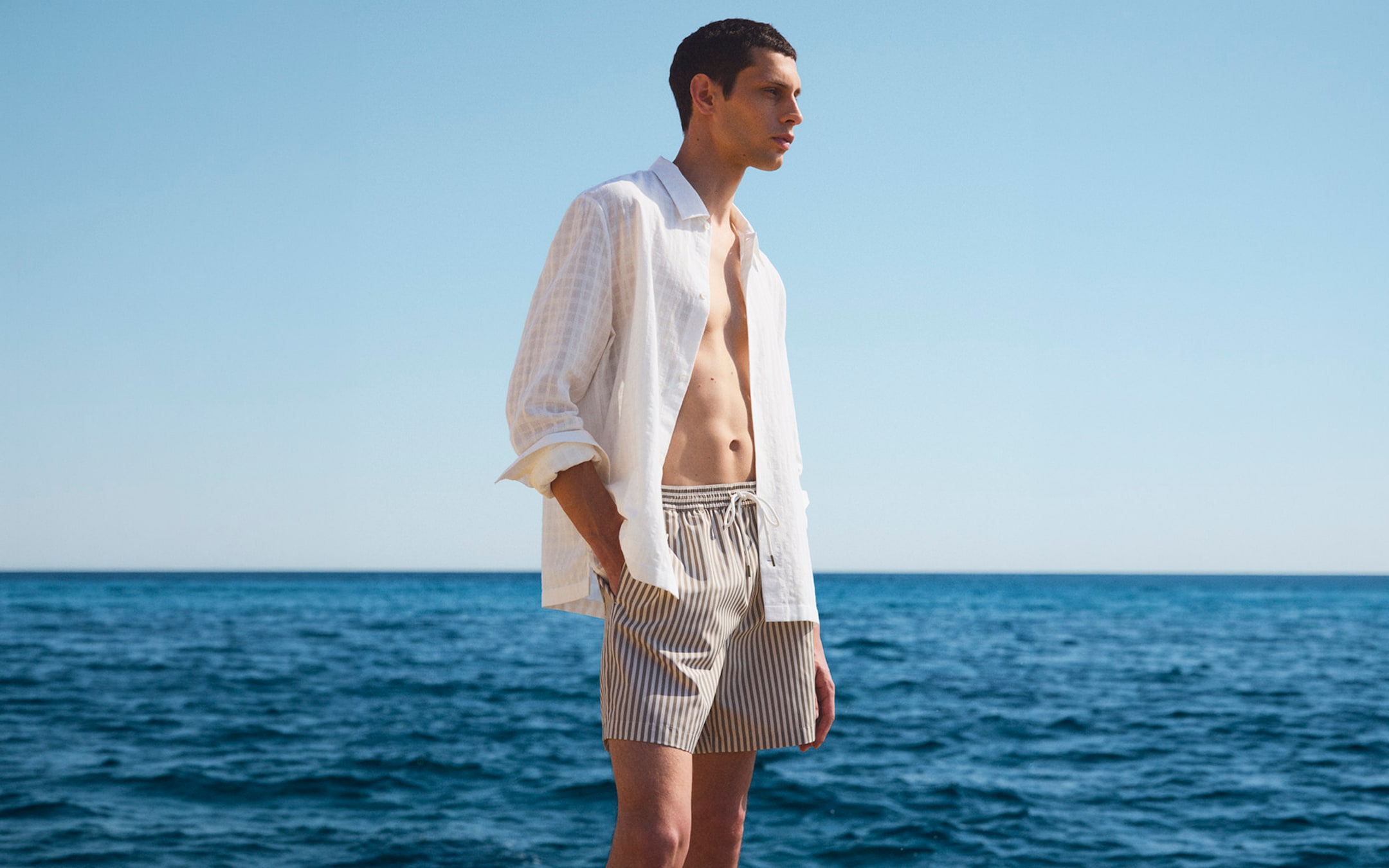 A person stands by the ocean wearing an open white shirt and striped shorts, with a clear blue sky in the background.