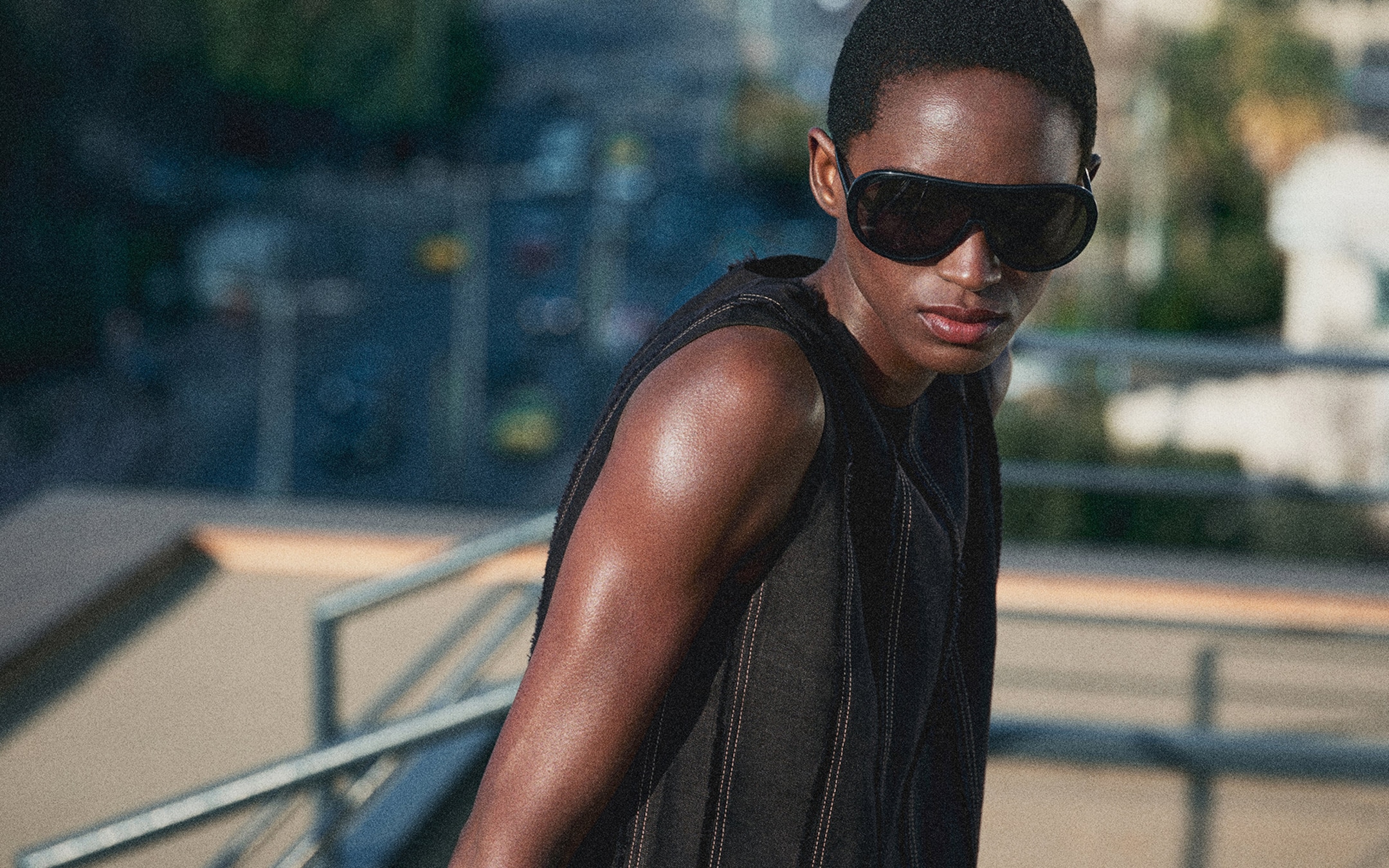 Person in a sleeveless outfit and large sunglasses, standing on a rooftop with a blurred urban background.