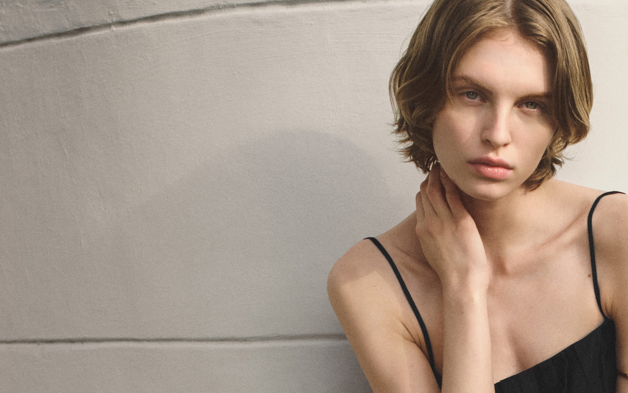 A person with short hair in a black spaghetti strap top poses against a light, curved wall, gently touching their neck.
