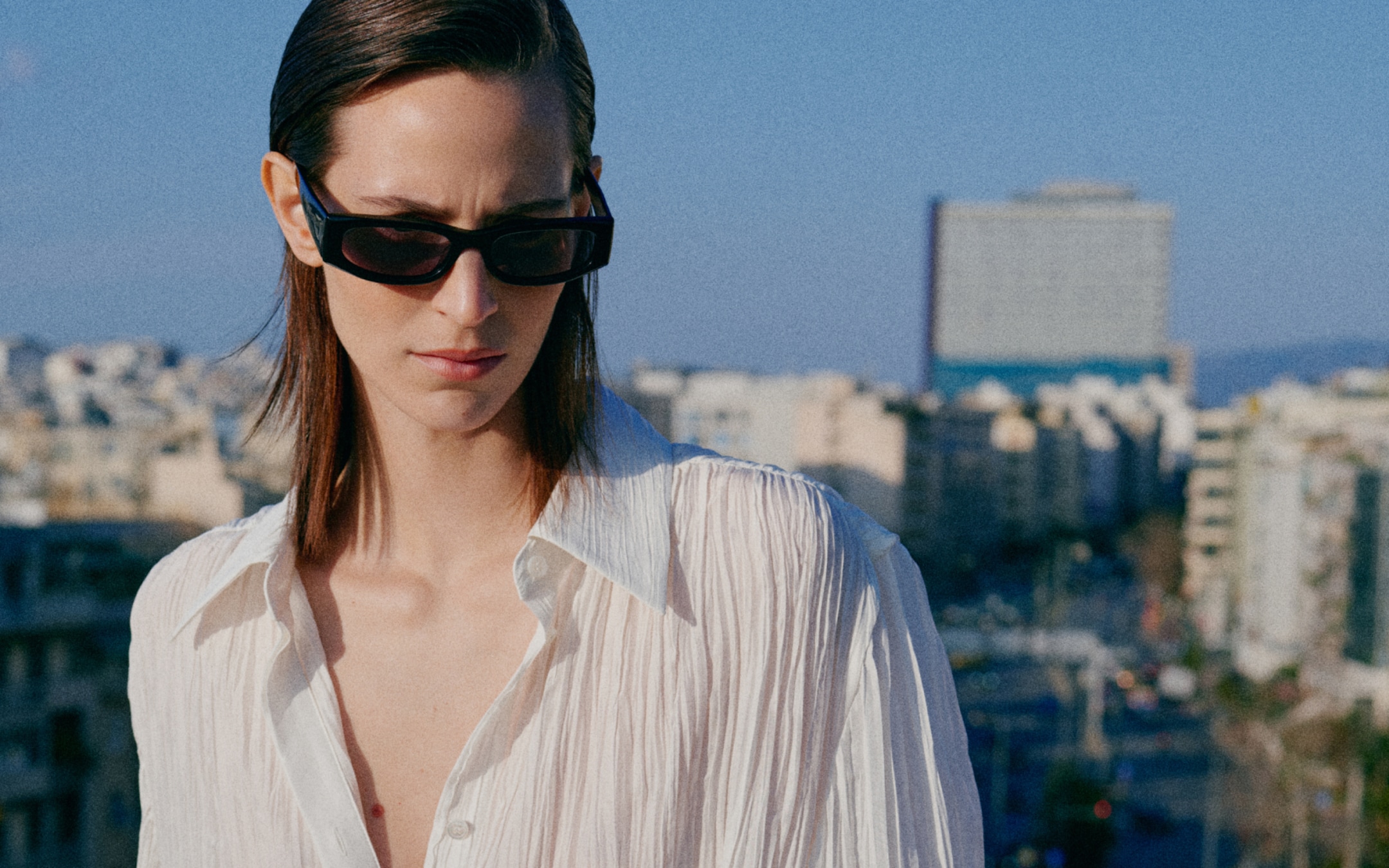 Person wearing sunglasses and a white shirt stands on a rooftop, with a cityscape and clear blue sky in the background.