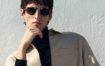 Placeholder for: Person wearing sunglasses and a light jacket, posing thoughtfully against a textured wall.
