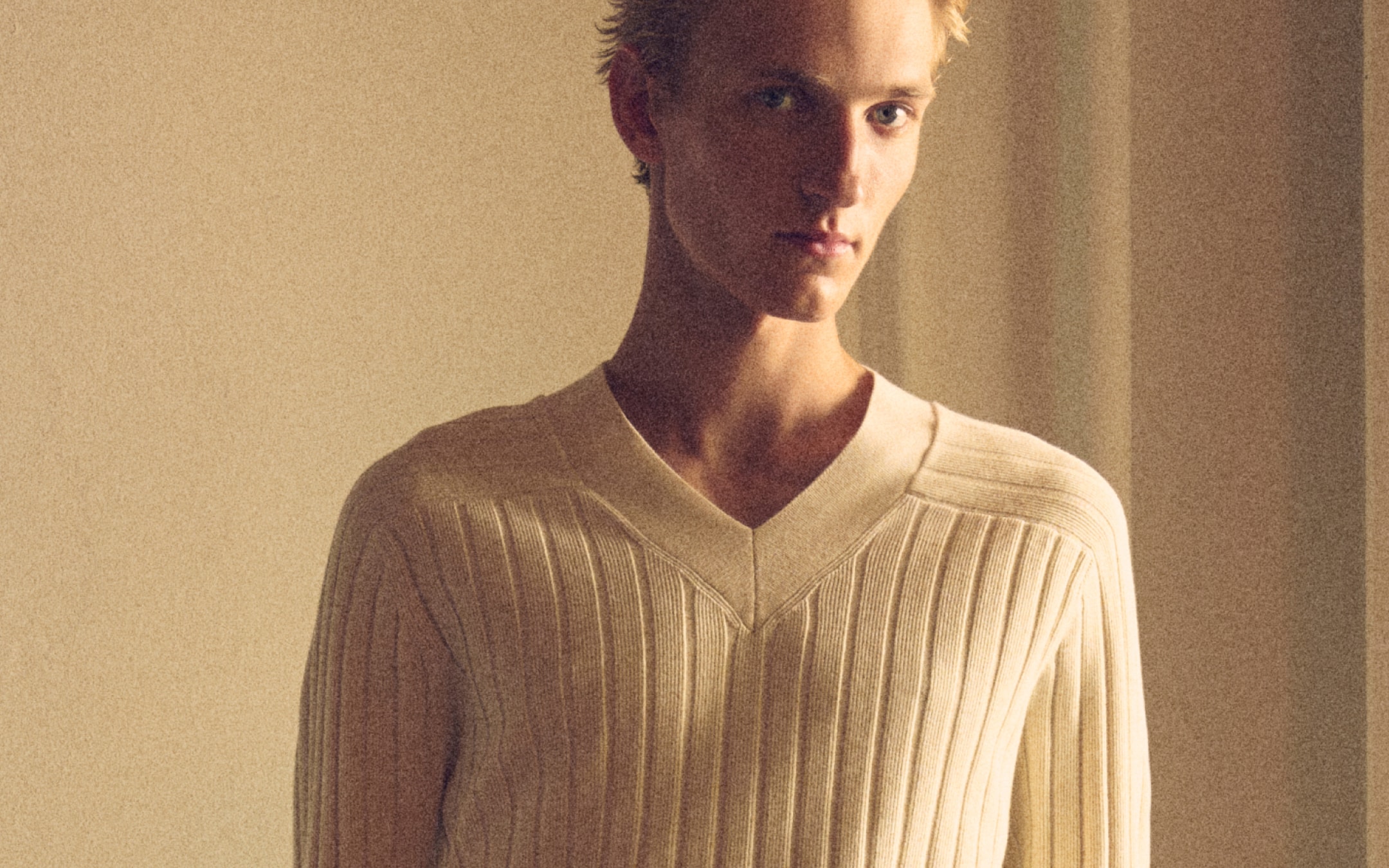 Close-up portrait of a blond young man in a cream ribbed V-neck sweater, soft warm lighting and neutral background.