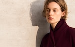 Placeholder for: Person with short hair wearing a dark maroon coat, standing against a textured wall, gazing thoughtfully to the side.