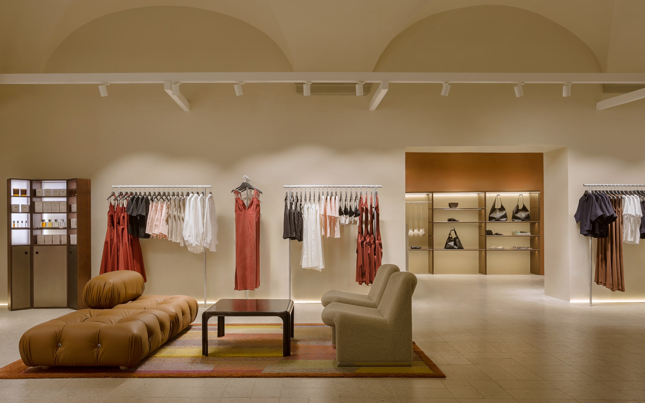 Chic clothing store with dresses on racks, cozy seating, and shelves displaying bags and accessories under warm lighting.
