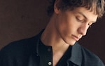 Placeholder for: A close-up of a young man with curly hair looking down, wearing a dark jacket over a green shirt, against a dark background.