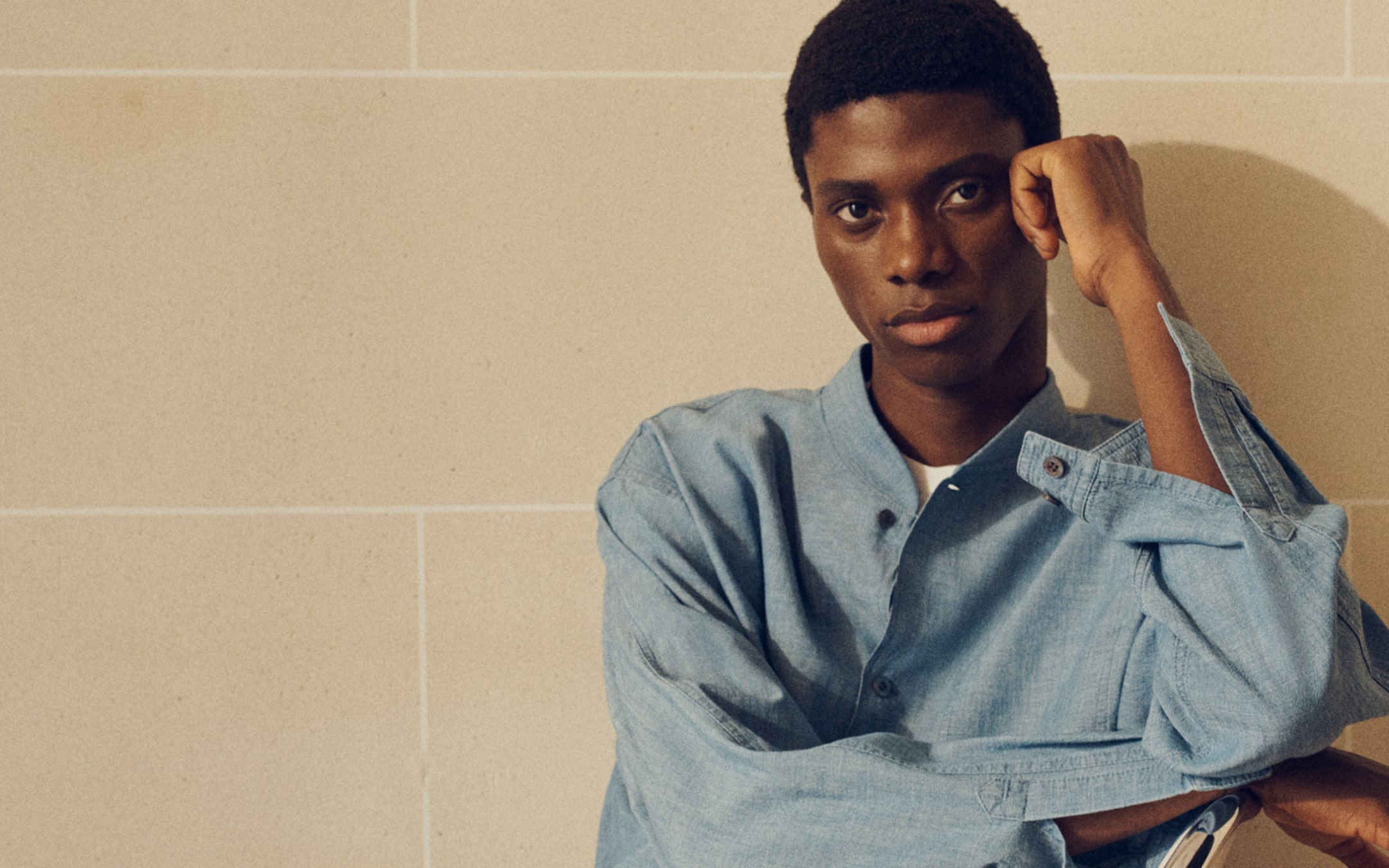 Person in a light blue denim shirt leaning against a beige tiled wall, gazing thoughtfully at the camera.