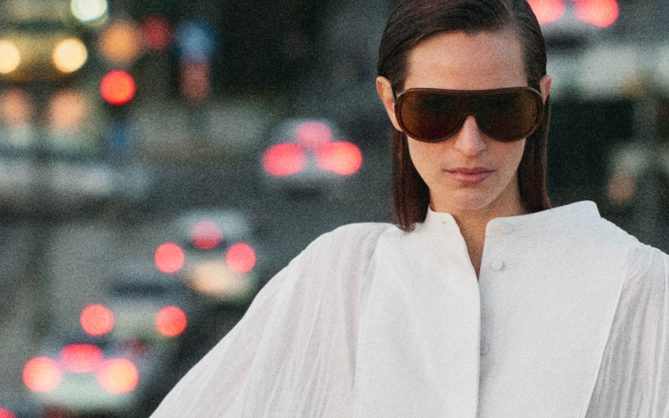 Woman with brown sunglasses and white blouse stands in front of a blurred urban background with city lights.