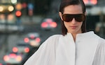 Placeholder for: Woman with brown sunglasses and white blouse stands in front of a blurred urban background with city lights.