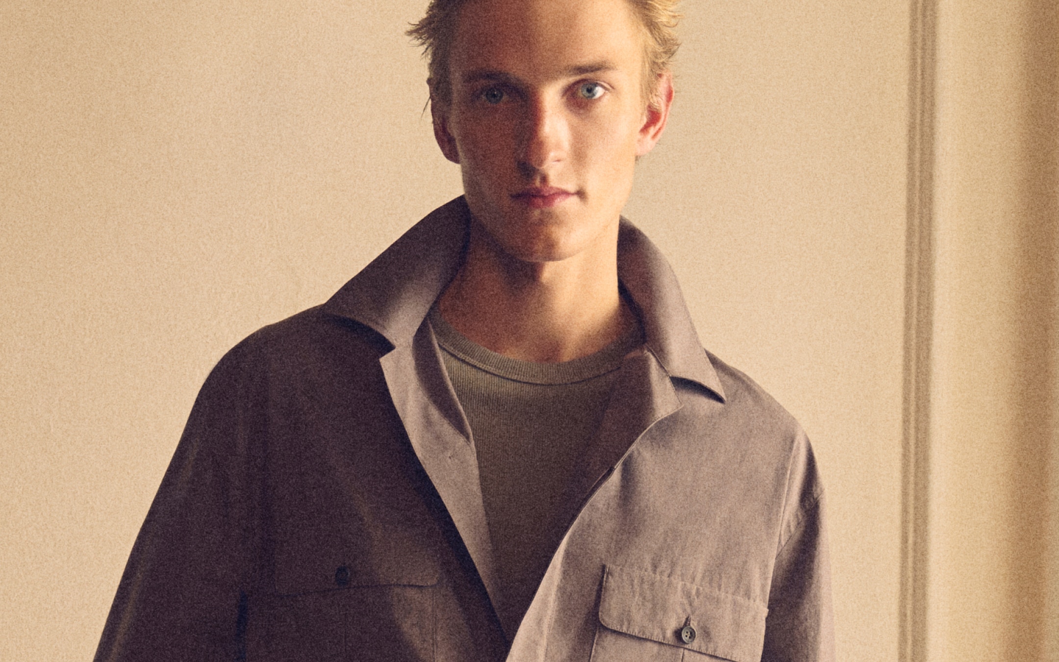 Close-up portrait of a young man in a collared jacket and knit shirt against a beige background, neutral expression.