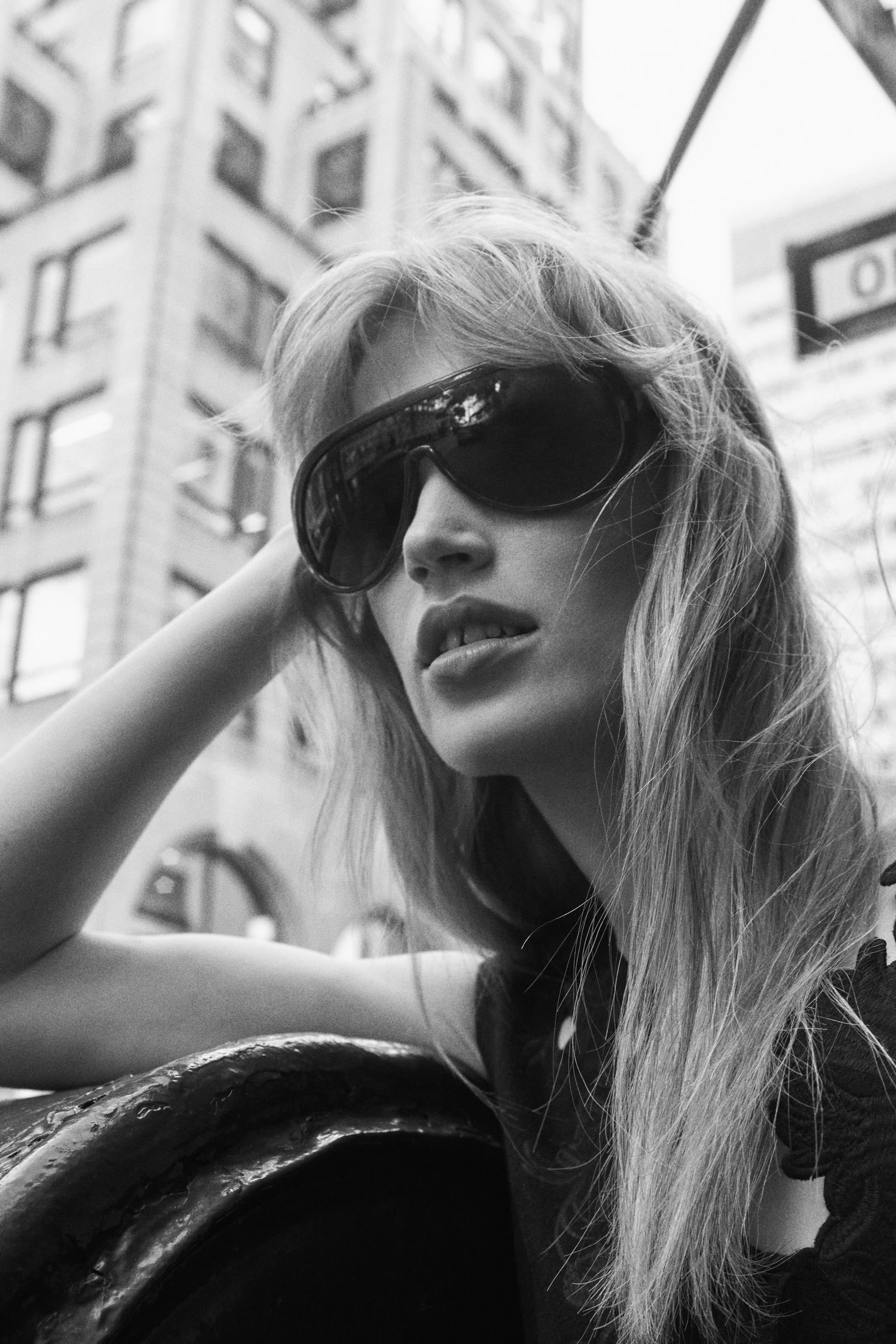 Black and white photo of a woman with long hair, wearing large sunglasses, looking thoughtfully into the distance against an urban backdrop.