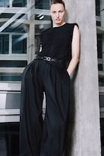 Placeholder for: Person in black outfit leans against a concrete pillar, wearing high-waisted pleated trousers and a textured top, in an urban setting.