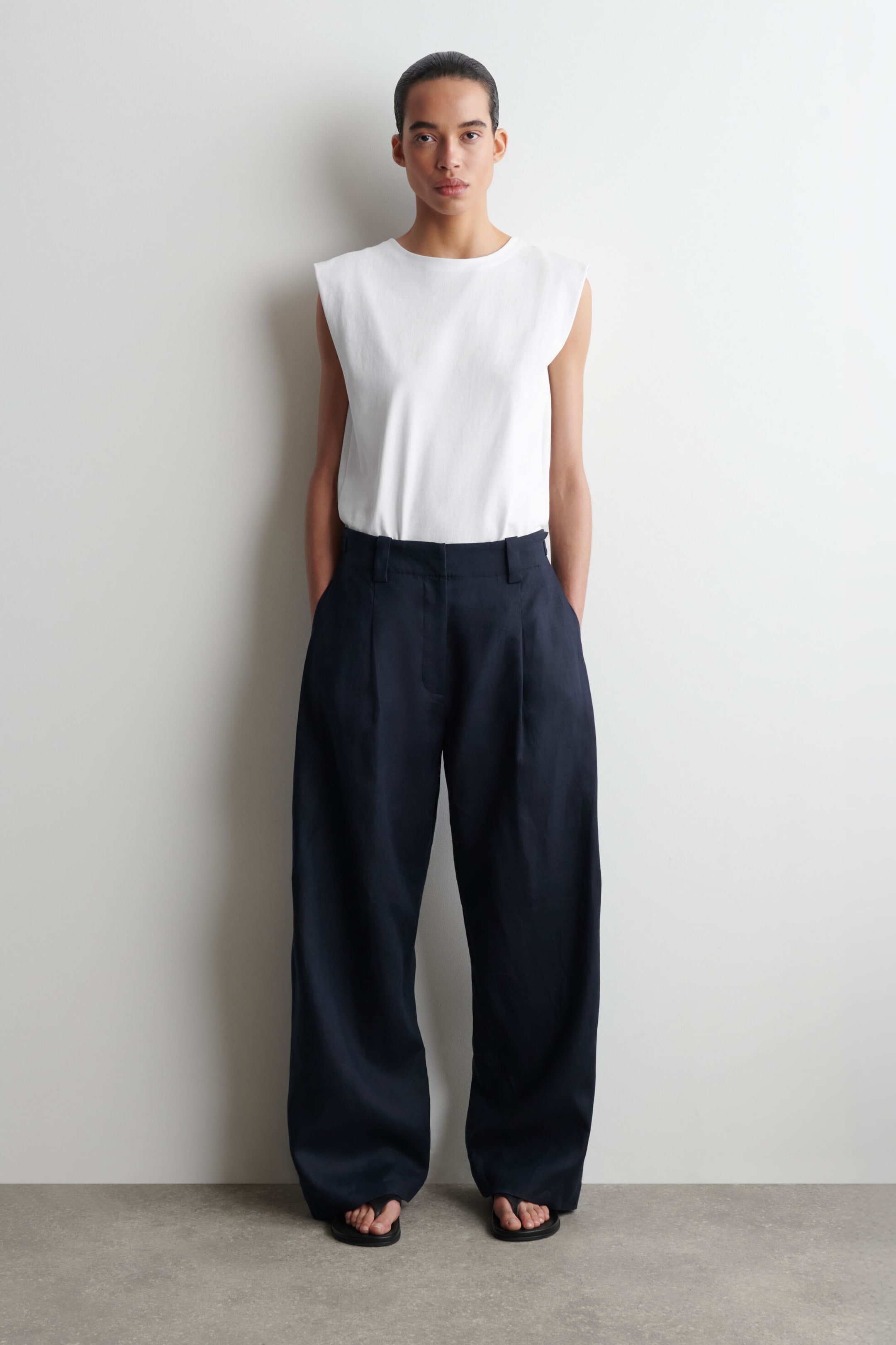 Person in a white sleeveless top, black wide-leg pants, and sandals, standing against a plain white wall.