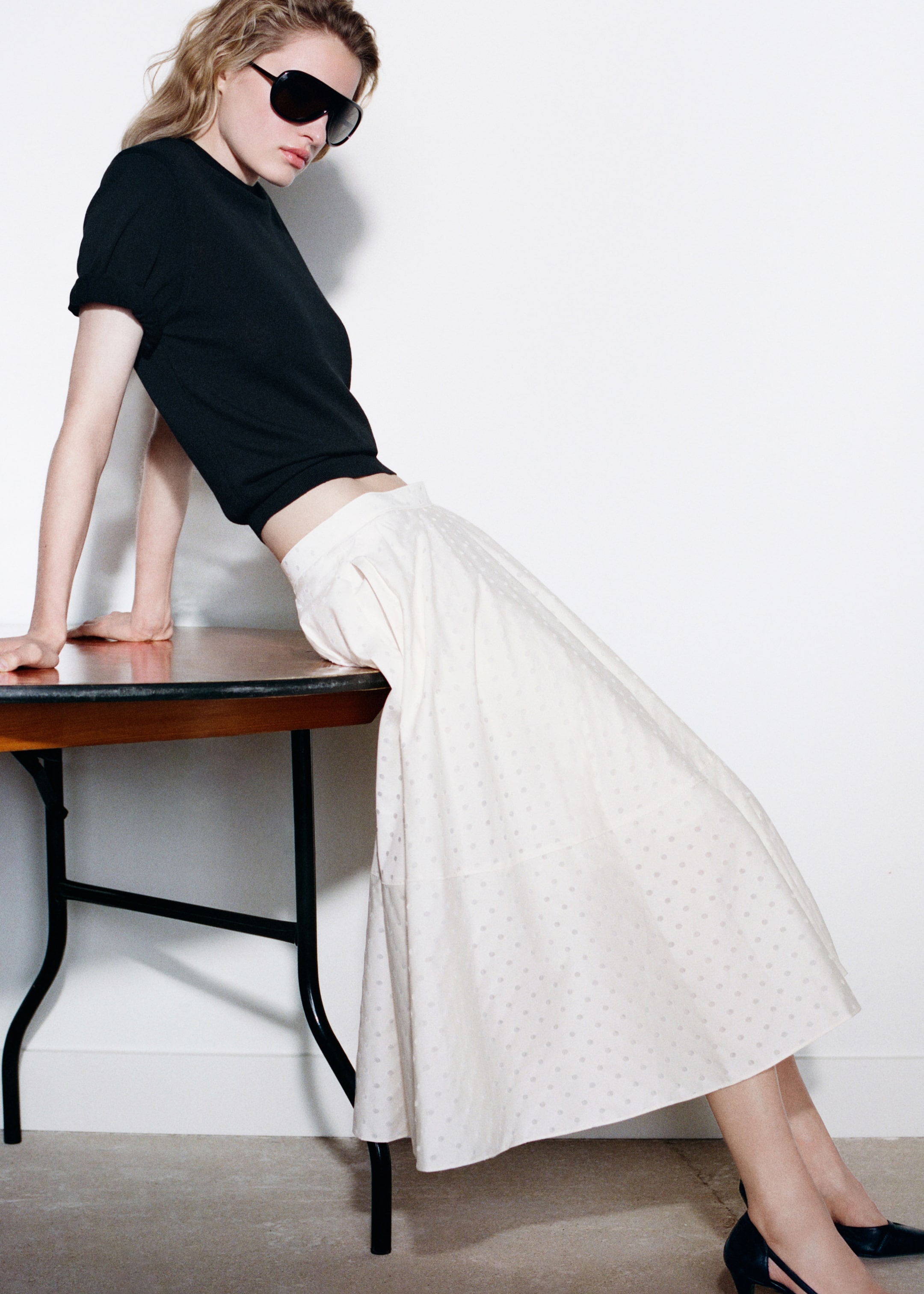 Woman in sunglasses leans against a table, wearing a black top and long white skirt, with black heels, in a minimalist setting.
