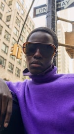 Placeholder for: Person wearing large sunglasses and a purple sweater, standing in an urban setting with a "One Way" sign visible.