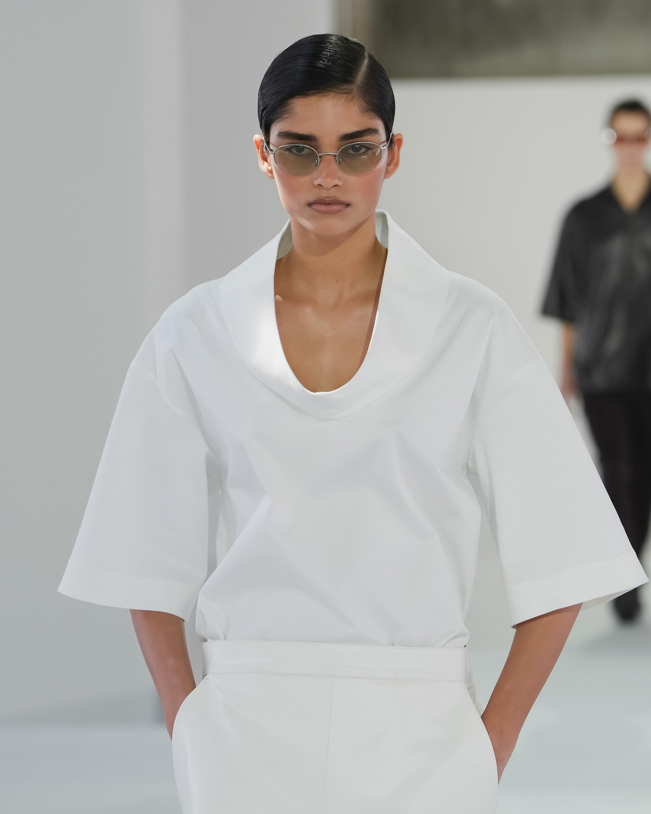 Model in white shirt with wide collar walks runway, hands in pockets, wearing sunglasses. Another model slightly blurred in the background.