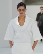 Placeholder for: Model in white shirt with wide collar walks runway, hands in pockets, wearing sunglasses. Another model slightly blurred in the background.