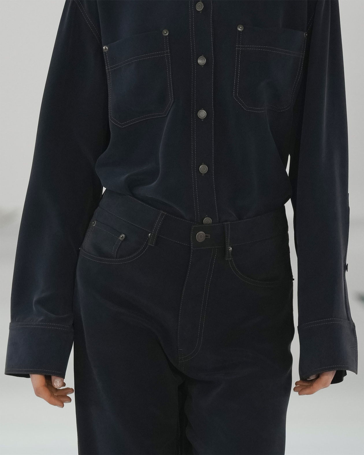 Person wearing a dark denim outfit with large cuffs, visible stitching, and metal buttons, standing against a light background.
