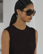 Placeholder for: Person wearing large sunglasses, a dark textured sleeveless top, and hair in a sleek ponytail. Neutral background.