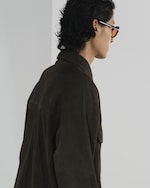 Placeholder for: Person with dark wavy hair wearing sunglasses and a brown jacket, captured from the side against a simple background.
