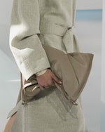 Placeholder for: Person in a beige outfit holding a large, soft, tan clutch bag. The coat has a subtle belt and long sleeves.