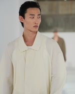 Placeholder for: Person in a light cream jacket with dark, slicked-back hair, looking to the side in a minimalist setting.