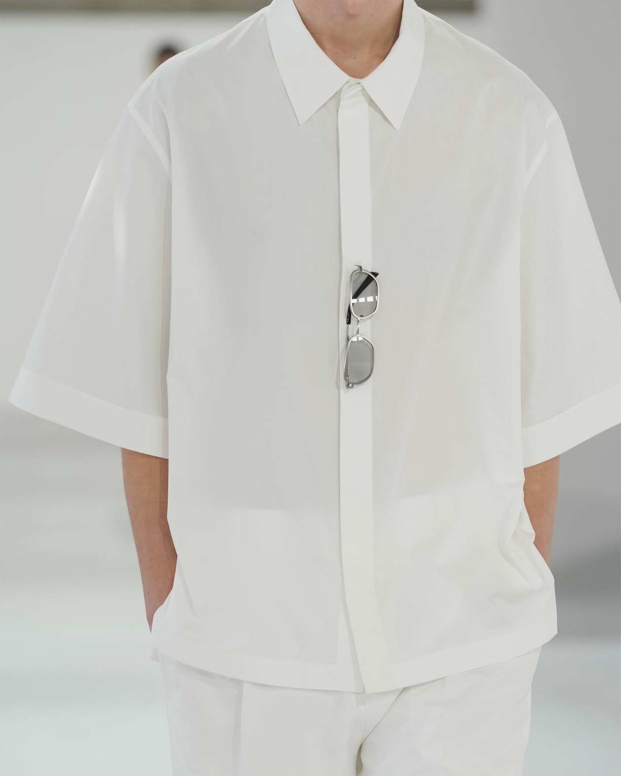 Person wearing a loose-fitting white shirt with sunglasses hanging from the placket. Their hands are in their pockets.