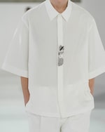 Placeholder for: Person wearing a loose-fitting white shirt with sunglasses hanging from the placket. Their hands are in their pockets.