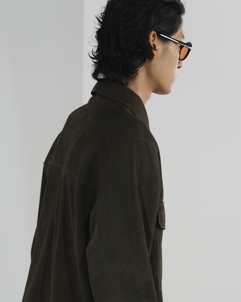 A person with dark hair wearing sunglasses and a brown suede jacket, shown from the side against a plain background.