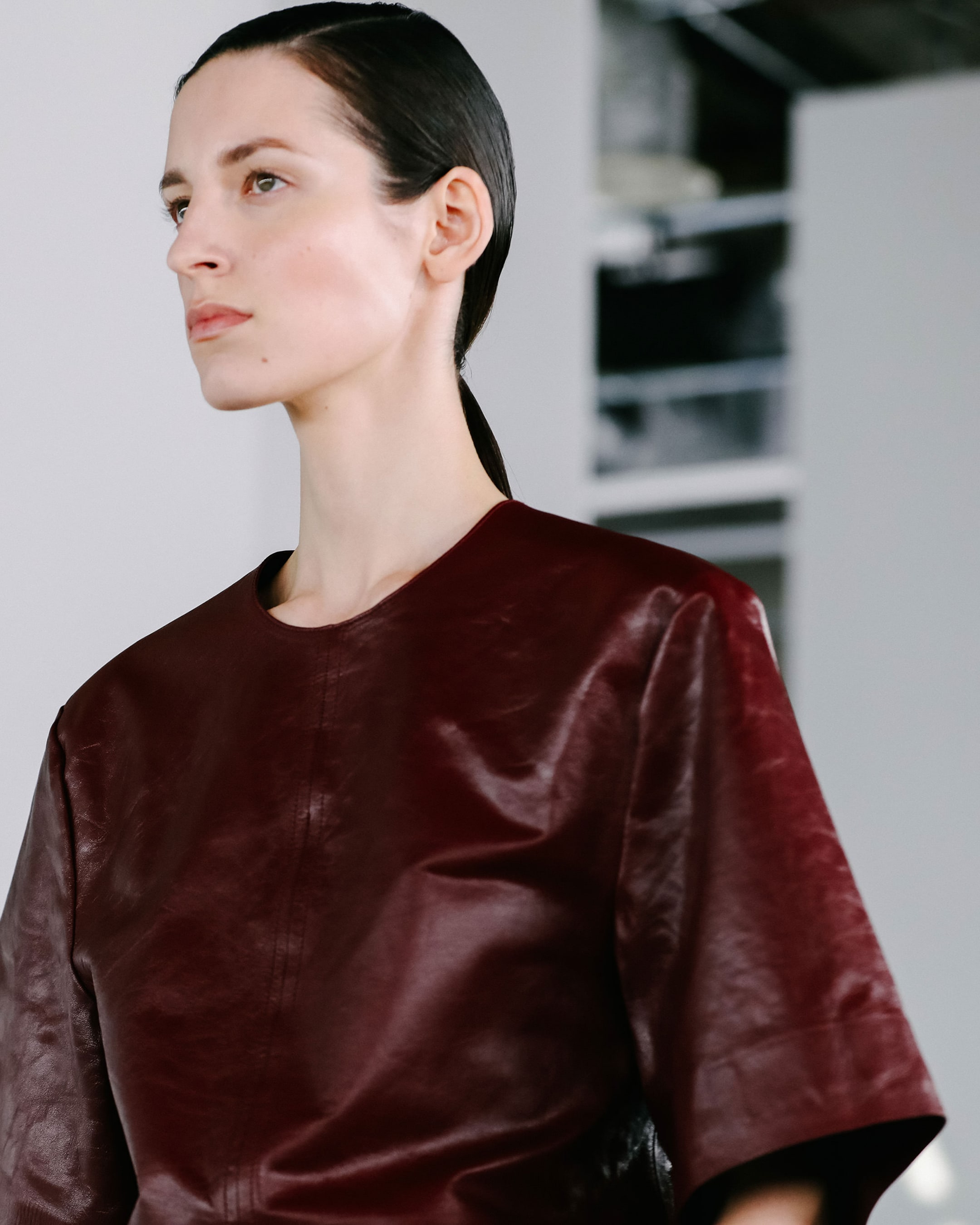Person in a sleek, dark red leather top with short sleeves and a ponytail, against a modern, minimalistic background.