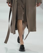 Placeholder for: Model wearing a long brown coat with shorts and black loafers, walking on a white runway.