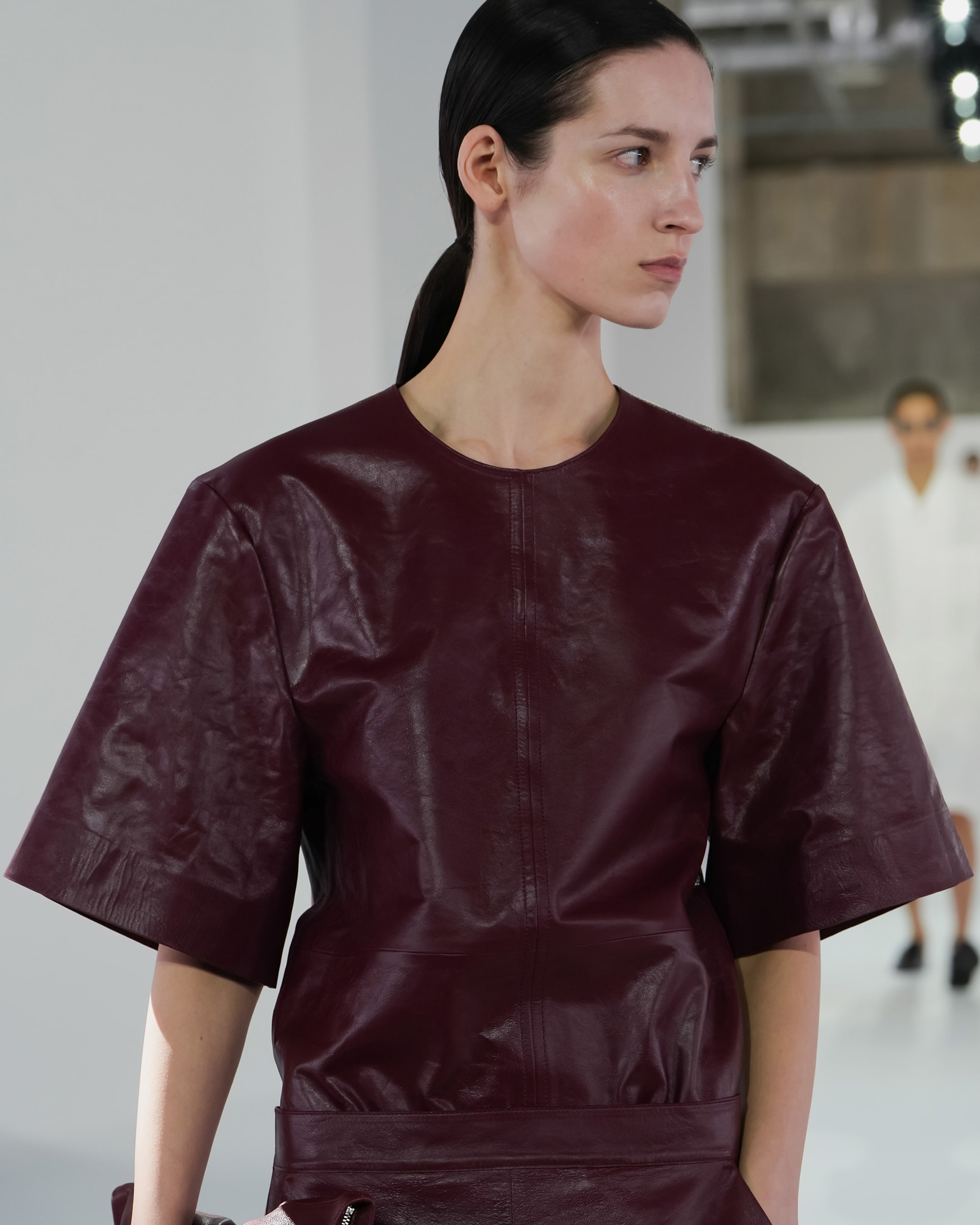A model wearing a maroon leather top with short, wide sleeves walks down a runway.
