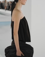 Placeholder for: A woman in a strapless black top holds a large black clutch, walking on a runway with blurred spectators in the background.