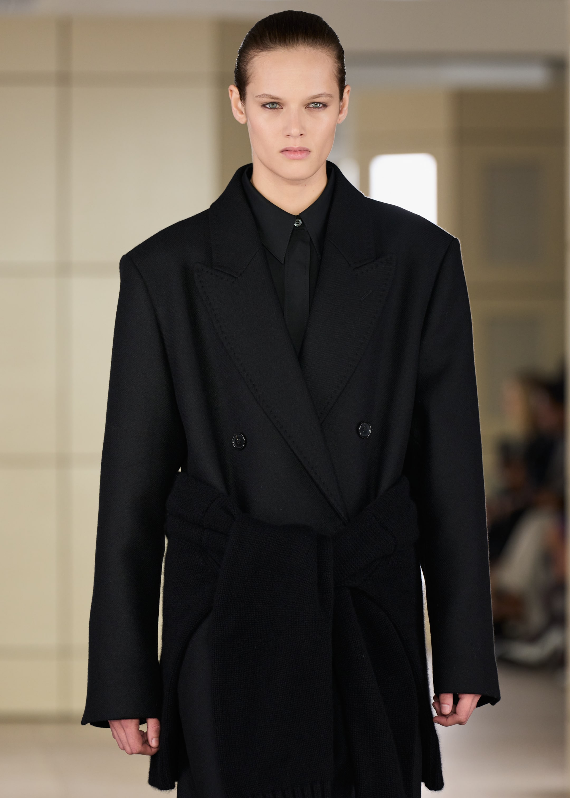 Model wearing a black tailored coat with a wide lapel and matching shirt, walking on a fashion runway with a blurred background.