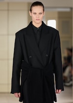 Placeholder for: Model wearing a black tailored coat with a wide lapel and matching shirt, walking on a fashion runway with a blurred background.
