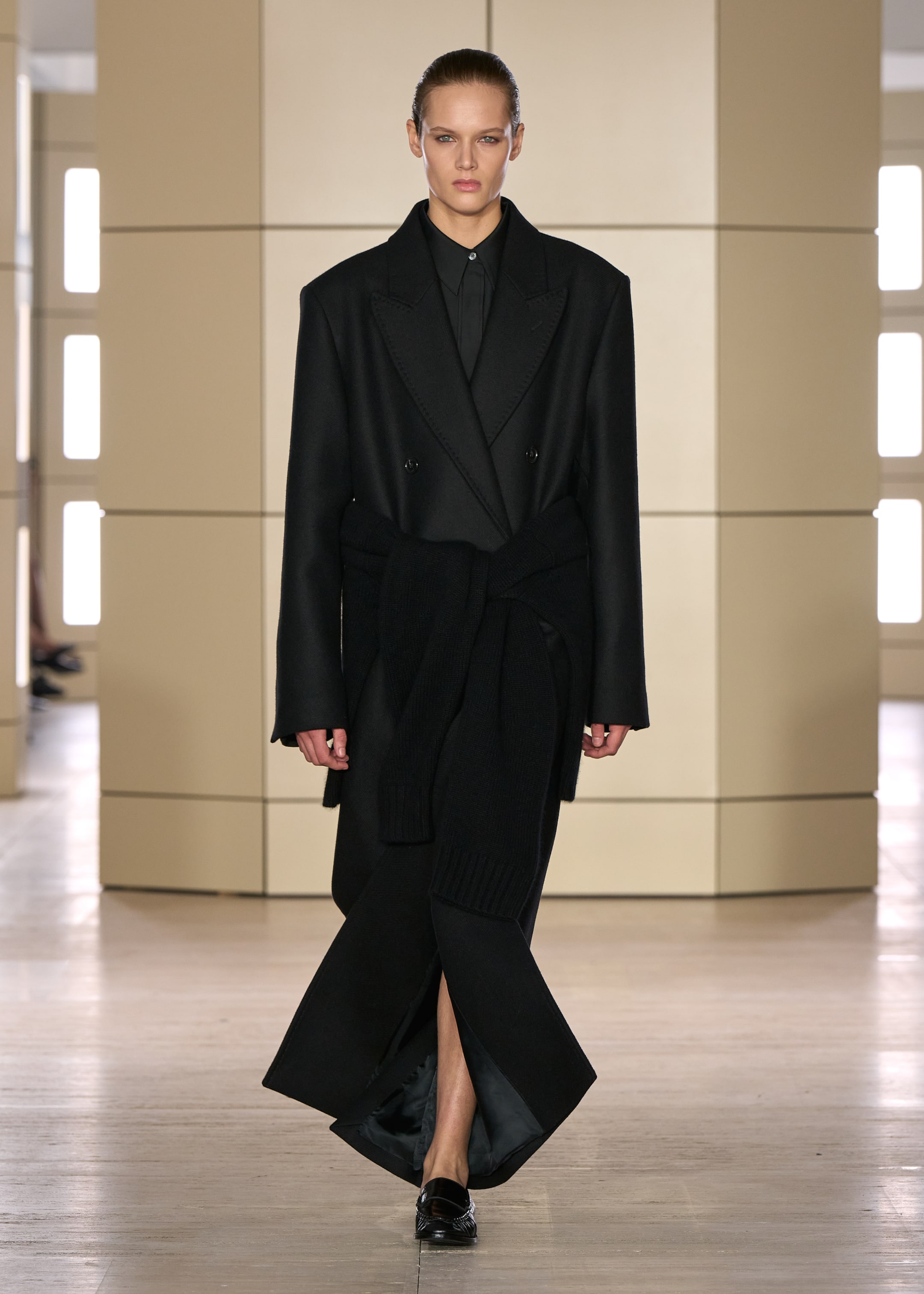 Model in an oversized black coat and long black skirt with a slit, walking on a runway with a minimalist beige background.