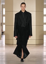 Placeholder for: Model in an oversized black coat and long black skirt with a slit, walking on a runway with a minimalist beige background.