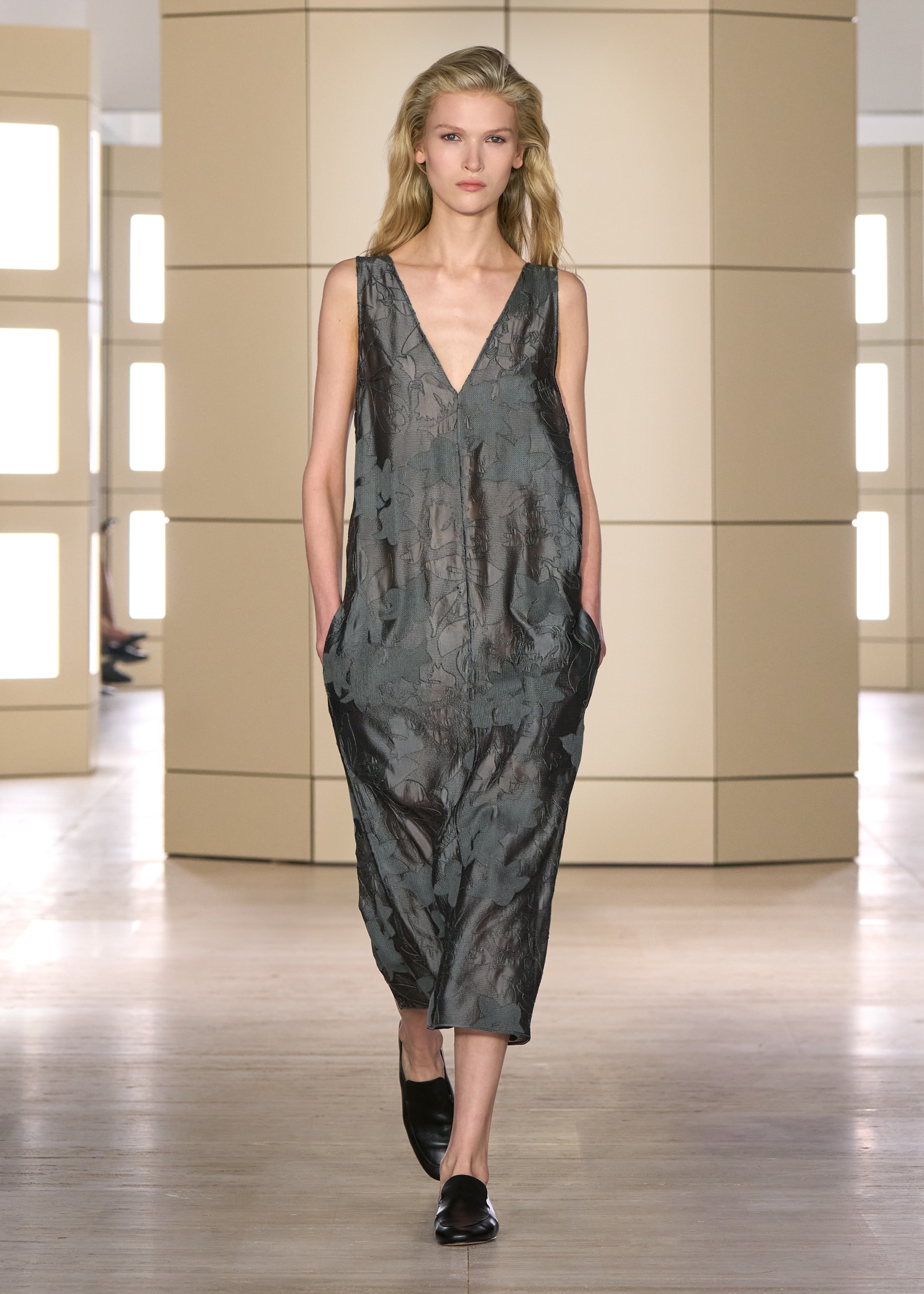 A model walks down a runway wearing a sleeveless, deep V-neck dress with a floral pattern and carrying her hands in the pockets.