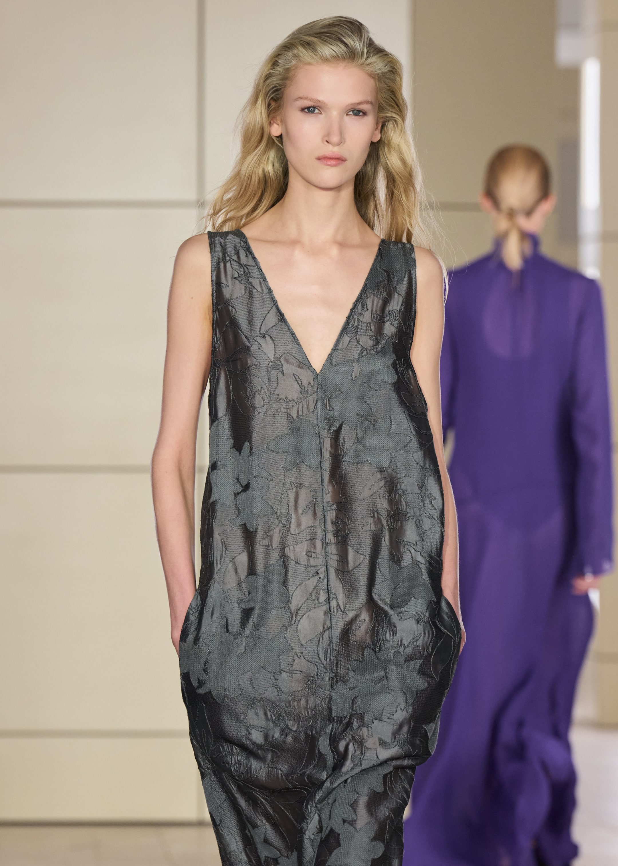 A model in a sleeveless, floral-patterned, dark gray dress walks down a runway. Another model in a purple outfit is seen in the background.