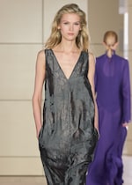 Placeholder for: A model in a sleeveless, floral-patterned, dark gray dress walks down a runway. Another model in a purple outfit is seen in the background.